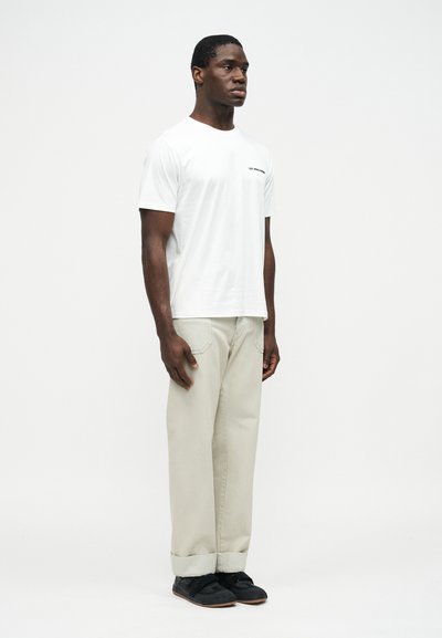 Άνδρας όρθιος, φοράει λευκό κοντομάνικο t-shirt με μικρό κείμενο στο στήθος, ανοιχτό μπεζ παντελόνι με ρεβέρ και μαύρα casual παπούτσια, μπροστά σε απλό φόντο.