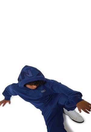 Conjunto de sudadera con capucha y pantalones en azul marino de tejido suave, con un gran logo en la capucha y un pequeño logo en el pecho. Zapatillas blancas.
