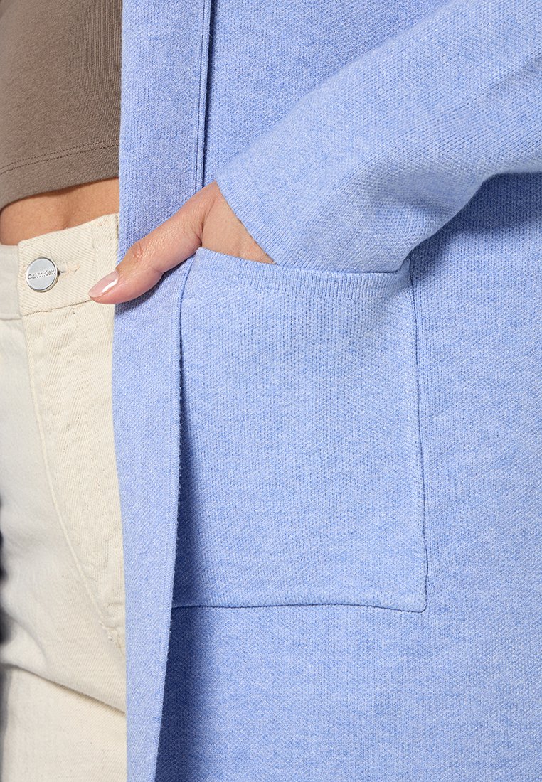 Hand mit manikürten Nägeln in der vorderen Tasche eines weichen, hellblauen Cardigans, der über beigen Hosen und einem braunen Oberteil getragen wird.