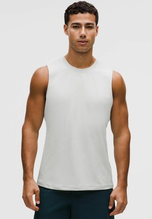 Fitte man met kort golvend haar, gekleed in een mouwloos wit sportshirt en donkere korte broek, staand tegen een effen achtergrond.