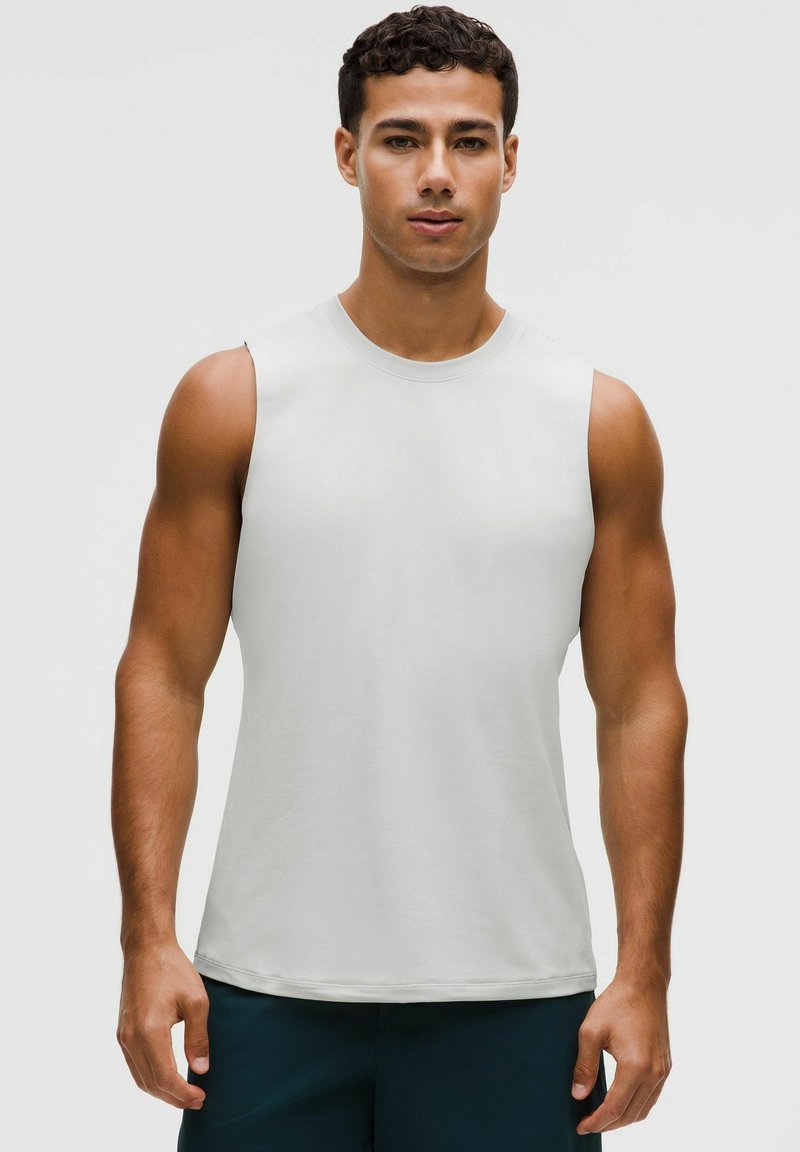 Hombre en forma con cabello corto y rizado, vistiendo una camiseta deportiva blanca sin mangas y pantalones cortos oscuros, de pie frente a un fondo liso.