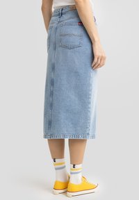 Jupe midi en denim bleu clair avec une coupe droite, coutures contrastées et deux poches arrière. Portée avec des baskets jaunes et des chaussettes rayées.