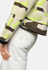 Pullover mit beigem und grünem Camouflage-Muster, braunen Akzenten und geripptem schwarzem Saum; kombiniert mit weißen Jeans mit detaillierter Naht.