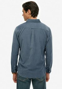 Uomo con camicia blu a maniche lunghe e jeans che guarda verso l'orizzonte su uno sfondo bianco semplice.