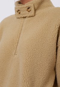 Beige fleece pullover met een hoge kraag, voorzien van een gedeeltelijke ritssluiting aan de voorkant en twee decoratieve knopen op de kraag. Zachte, gestructureerde oppervlakte.