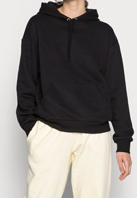 Zwarte hoodie van zachte katoen, met een ruime vozakker, brede manchetten en een verstelbare trekkoordkap. Gecombineerd met lichte broeken.