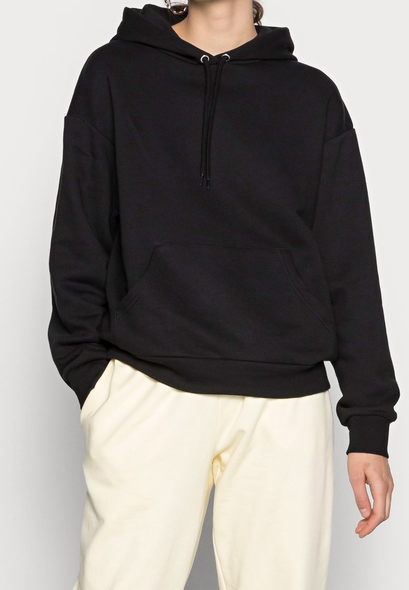 Zwarte hoodie van zachte katoen, met een ruime vozakker, brede manchetten en een verstelbare trekkoordkap. Gecombineerd met lichte broeken.