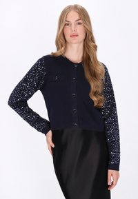 Sötétkék kardigán, testhezálló szabással, gombos elülső résszel és sima anyaggal. A hüvelyek flitter mintával rendelkeznek, ami textúrát és csillogást ad.