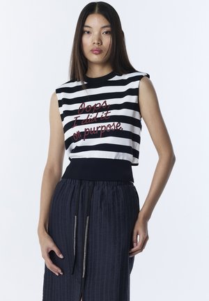 Giovane donna con lunghi capelli scuri indossa una canottiera a righe bianco e nere con la scritta "Oops, l'ho fatto apposta" e una gonna scura.