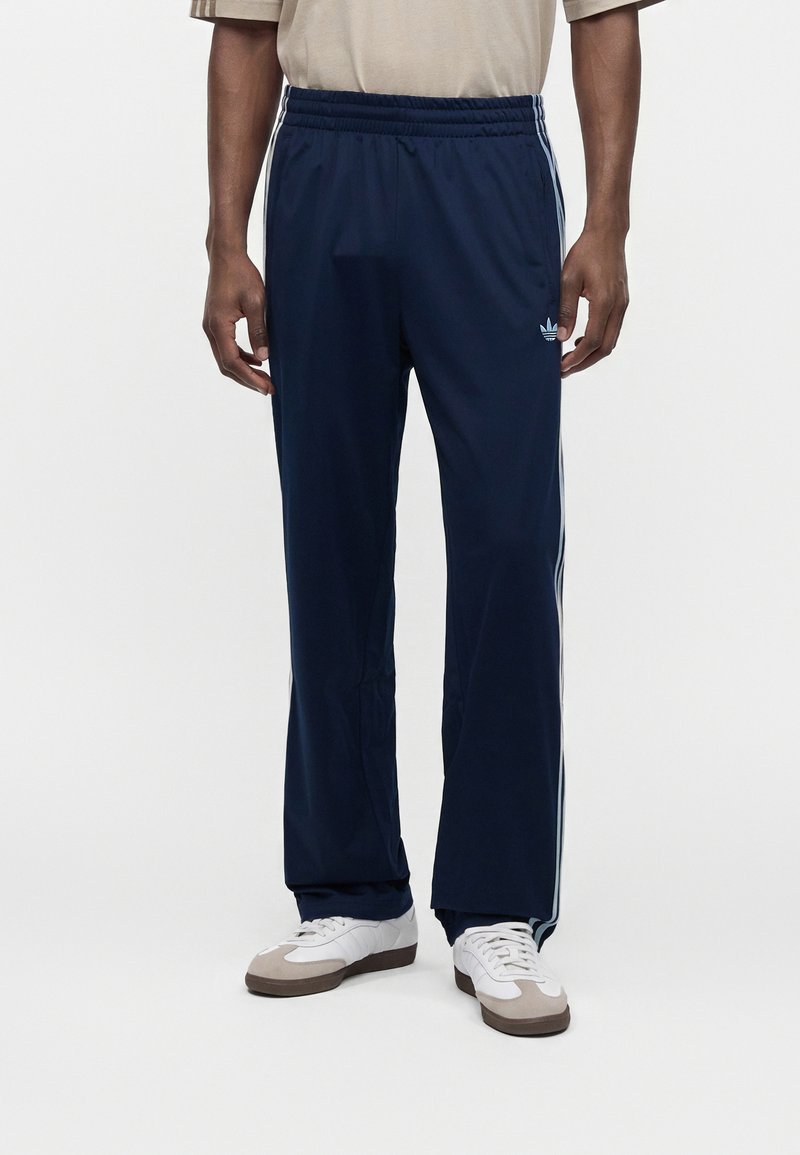 Man wearing navy blue Adidas track pants with white side stripes and white sneakers, standing against a plain white background.