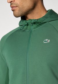 Zelena zip-up dukserica s teksturiranim kapuljačom, kontrastnim ramenskim panelima i malim brendiranim logotipom na prsima. Glatka tkanina, ležeran dizajn.