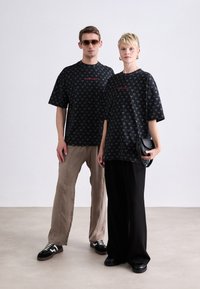 Sorte oversized t-shirts har et gentaget mønster og et rødt logo. Den mandlige model bærer beige bukser og sneakers; den kvindelige model holder en sort taske.