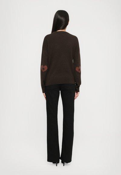 Woman with long dark hair stands facing away, wearing a dark brown sweater with heart patches on elbows and black flared pants.