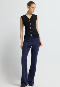 Czarny bezrękawnik z dekoltem w serek i pięcioma złotymi guzikami, zestawiony z ciemnonawiwowymi spodniami dzwonami. Gładki materiał, dopasowany krój i proste linie.