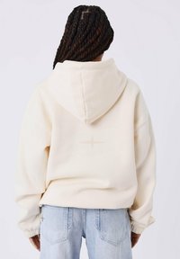 Persoon met donker gevlochten haar, gekleed in een crèmekleurige hoodie en lichtblauwe jeans, staand met de rug naar de camera tegen een effen witte achtergrond.