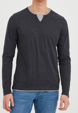 Man wearing a dark gray long-sleeve shirt with a gray undershirt peeking through the collar and light blue jeans, standing against a white background.