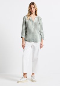 Camicia di lino a righe verde chiaro con scollo a V e maniche arrotolate, abbinata a pantaloni bianchi a gamba larga e sneakers chiare.