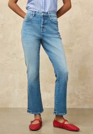 Person wearing light blue high-waist jeans and red woven Mary Jane flats, standing against beige textured wall.