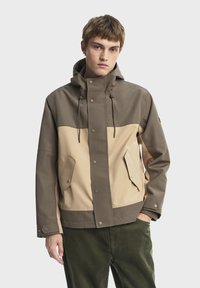 Hnedo-bežový dvojtonový kabát s kapucňou, patentnými gombíkmi a bočnými vreckami. Textúrovaná látka s matným povrchom. Styling so zelenými nohavicami.