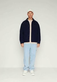 Jachetă albastră închis cu nasturi peste o cămașă bej deschis, asortată cu blugi albaștri deschiși și adidași albi. Modelul poartă o șapcă albă.