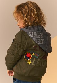 Veste bomber vert olive avec capuche grise, ornée de divers patchs et broderies. Tissu texturé, poignets côtelés et poche zippée.