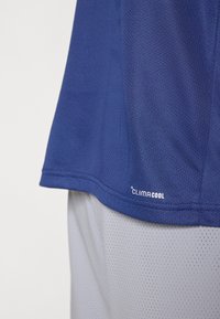 Μπλε ναυτικό αθλητικό πουκάμισο από διαπνέον ύφασμα με υφή από διχτυωτό ύφασμα· διαθέτει λογότυπο "CLIMACOOL" στο κάτω μέρος του. Συνδυάζεται με ανοιχτό γκρι σορτς.