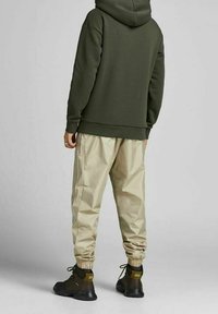 Sudadera verde oliva con capucha y cuerda ajustable, combinada con joggers beige ajustados que presentan puños elásticos y una franja lateral. Calzado negro.