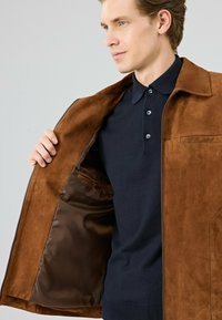 Bruine suède jas met een ritssluiting, voorzien van een zachte voering en een borstzak. Draagt over een donkere polo. 