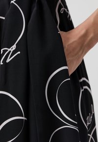 Crna tkanina s bijelim kružnim uzorcima i natpisom, koja pokazuje glatku teksturu. Ima bočnu džepu i elegantan pad.