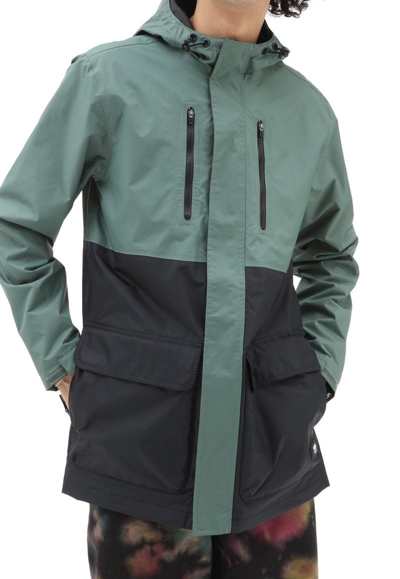 Homme portant une veste à capuche verte et noire avec des poches poitrine zippées et de grandes poches à rabat, mains dans les poches, pantalon multicolore visible.