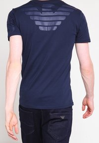 Námořnická modrá tričko s horizontálním pruhovaným designem na zadní straně, vyrobené z měkké tkaniny. Úzký střih, spárováno s tmavě modrými džíny s kapsami.