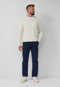 Mann steht vor einem hellgrauen Hintergrund, trägt einen cremefarbenen Pullover, marineblaue Hose und weiße Sneaker und schaut nach links.