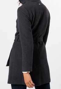 Abrigo de lana gris oscuro con cintura ceñida, mangas largas y bolsillos laterales inclinados. Presenta un cuello con muesca y un discreto detalle de logo en la parte posterior.