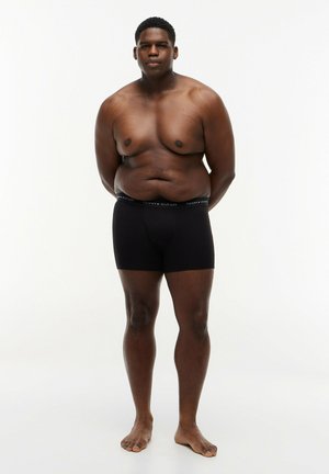 Uomo dalla pelle scura in piedi a piedi nudi, indossa boxer neri, con le braccia dietro la schiena, rivolto verso avanti su sfondo bianco uniforme.