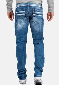 Jeans in denim blu con vestibilità slim, caratterizzati da tasche laterali con cerniera, sottili scoloriture e accenti di cucitura. Indossati con sneakers bianche.