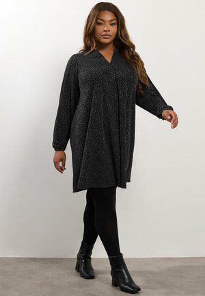 Vestido negro con un sutil patrón texturizado, escote en V, mangas largas y un diseño fluido, combinado con leggins negros y botas de tobillo.