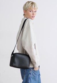 Bolso cruzado de cuero negro con forma rectangular, que presenta un zipper plateado y un logo sutil. Llevado sobre un suéter de color claro.