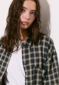 Cămașă cu carouri din flanel în nuanțe de navy și crem, cu guler cu nasturi, croială lejeră și un buzunar frontal. Purtată peste un tricou alb.