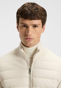 Helle beige gepolsterte Jacke mit hohem Kragen, durchgehendem Reißverschluss und gerippten Akzenten. Getragen über einem passenden Rollkragenpullover. Glatte Textur.