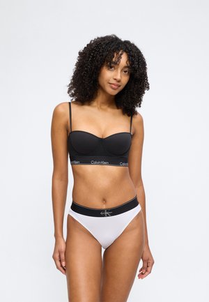 Femme aux cheveux bouclés portant un bralette noir Calvin Klein et une culotte taille haute blanche Calvin Klein, debout devant un fond uni.