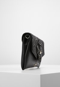 Sac à main noir avec un rabat texturé et scintillant, comprenant une décoration de fleur noire accentuée et des ferrures métalliques. Corps en cuir lisse.