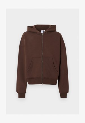 Młody mężczyzna o czarnych włosach, ubrany w brązową bluzę na zamek z kapturem i pasujące spodnie, stojący na tle jednolitego jasnego tła.