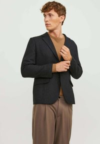 Blazer nero in tessuto texturizzato con revers a punta, chiusura a un bottone e due tasche, abbinato a un maglione marrone e pantaloni beige.