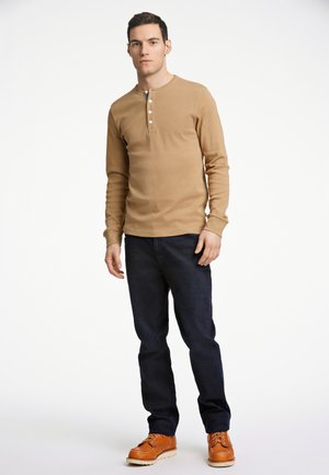 Langärmliges, beiges Henley-Shirt mit drei Knöpfen, kombiniert mit dunklen Jeans und braunen Schnürstiefeln. Glatte Textur und taillierte Passform.