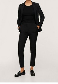 Costume noir comprenant un blazer ajusté et un pantalon cintré, tous deux en tissu élégant. Associé à des mocassins noirs brillants, créant un look cohérent.