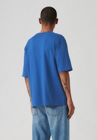 Μπλε κοντομάνικο μπλουζάκι από βαμβάκι, με χαλαρή εφαρμογή, στρογγυλή λαιμόκοψη και ορατές ραφές. Φορεμένο με ανοιχτό μπλε τζιν.