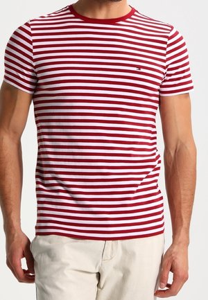 Mand iført en rød og hvid kortærmet T-shirt med horisontale striber og beige bukser mod en ensfarvet baggrund.