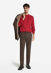 Röd ulltröja, bruna byxor och svarta snörskor. Håller en brun shearling-fodrad kappa. Enkel design och passform.