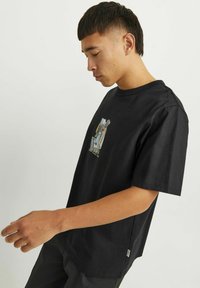 Camiseta de algodón negra de manga corta, con un diseño gráfico en la parte delantera. El modelo la lleva puesta, vista de lado.