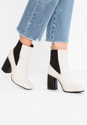 Bottes de cheville blanches et noires avec un talon bloc haut, présentant une finition en cuir lisse et des panneaux latéraux élastiques. Un jean en denim déchiré complète le look.